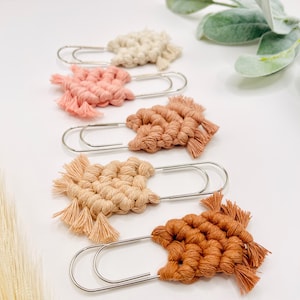May include: Five large silver metal paper clips, each adorned with a unique macrame knot design in varying shades of cream, light pink, dusty rose, beige, and terracotta. Each macrame piece features a fringed edge and is securely attached.