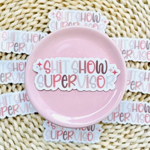 Pu&ograve; includere: Un adesivo rosa e bianco con il testo "SHIT SHOW SUPERVISOR" in un font colorato e stratificato. L'adesivo &egrave; su un piatto rosa e circondato da altri adesivi con lo stesso testo.