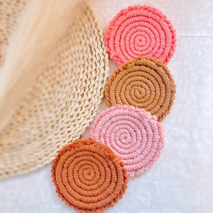 May include: Four round crocheted coasters in shades of pink and brown. The coasters have a fringed edge and a spiral design.