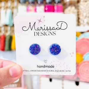 May include: A pair of blue glitter stud earrings with pink glitter accents. The earrings are displayed on a white card with the text "Marissa D DESIGNS" and "handmade" on it.