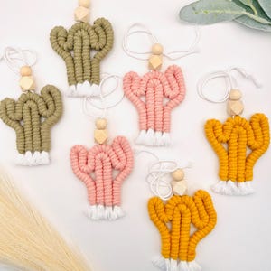May include: Six handmade macrame cactus ornaments with wooden beads and white fringe. The cacti are in various colors, including green, pink, and yellow.