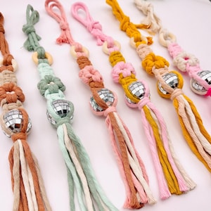 May include: A collection of colorful macrame keychains. Each keychain features a small disco ball, wooden ring, and knotted macrame design in shades of pink, yellow, brown, green, and cream. The tassels add a decorative touch.