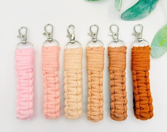 May include: Six macrame wristlet keychains arranged in a row, featuring a color gradient from light pink to dark orange. Each keychain is crafted with knotted cotton cord and includes a silver-toned metal lobster clasp at the top for attachment.