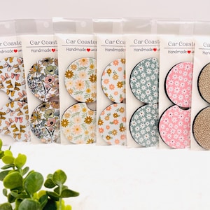 May include: A collection of handmade car coasters in various floral patterns. Designs include bows, daisies, and other flowers in colors like pink, yellow, and green. Each coaster is packaged in a clear plastic sleeve.