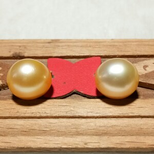 Puede incluir: Dos perlas redondas y lustrosas de tono dorado est&aacute;n colocadas una al lado de la otra. Las perlas est&aacute;n montadas sobre un peque&ntilde;o soporte rojo en forma de mariposa, fijado a una tira de madera horizontal de color marr&oacute;n claro.