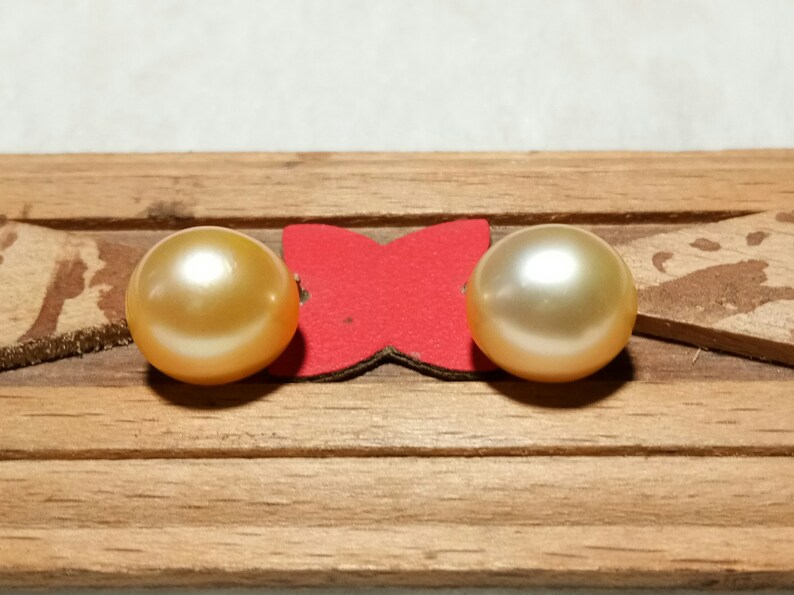 Puede incluir: Dos perlas redondas y lustrosas de tono dorado est&aacute;n colocadas una al lado de la otra. Las perlas est&aacute;n montadas sobre un peque&ntilde;o soporte rojo en forma de mariposa, fijado a una tira de madera horizontal de color marr&oacute;n claro.