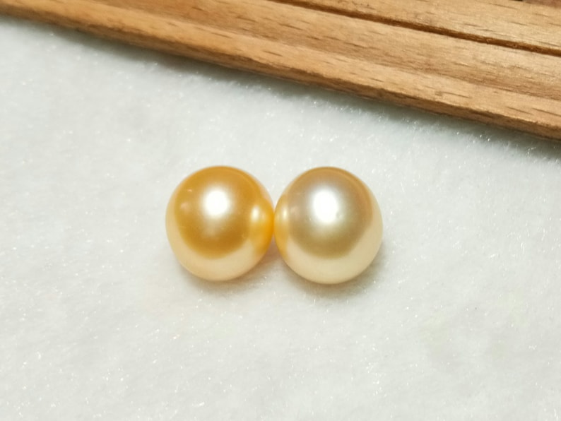 Puede incluir: Dos perlas redondas de tono dorado con una superficie lisa y brillante. Cada perla presenta un brillo reflectante y un color amarillo met&aacute;lico c&aacute;lido, mostrando una forma esf&eacute;rica y un acabado pulido que captura la luz de manera uniforme.