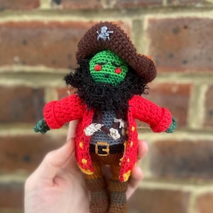 May include: A handmade crochet pirate doll with a green face, red eyes, and a black beard. The doll wears a brown pirate hat, a red coat with gold trim, and brown boots. The doll is approximately 6 inches tall.