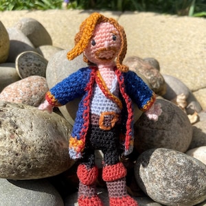 May include: A crocheted pirate doll with long, orange hair, a blue jacket with red trim, a white shirt, a black belt, black pants, and red boots.