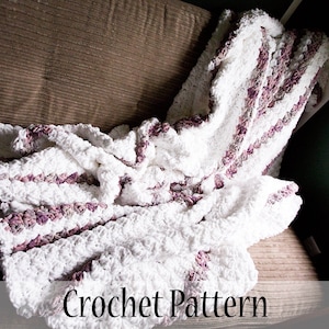 Może przedstawiać: Biały koc szydełkowy w pasy fioletowe i białe. Koc jest złożony i drapowany na brązowej kanapie. Tekst "Crochet Pattern" jest widoczny na dole obrazu.