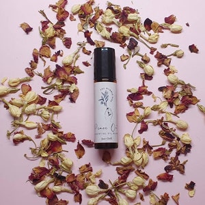 May include: A black essential oil roller bottle with the text "Peace Oil" and "Just Chill" on a white label. The bottle is surrounded by dried rose petals and white flower buds on a pink background.