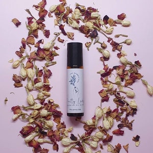 May include: A small glass bottle with a black roller ball top. The bottle is filled with a clear liquid and has a white label with the text "The Pretty Lady" and "Soothing Body Oil". The bottle is surrounded by dried rose petals and white flower buds on a pink background.