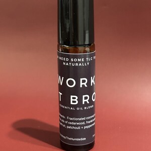 May include: A dark glass essential oil blend bottle with a black cap. The label reads "WORK IT BRO" and "ESSENTIAL OIL BLEND". The ingredients include fractionated coconut oil, cedarwood, frankincense, patchouli, and peppermint.
