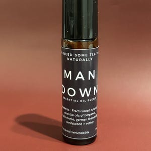 May include: A dark glass essential oil blend bottle with a black cap. The label reads "MAN DOWN ESSENTIAL OIL BLEND" with a list of ingredients. The bottle is on a reddish-brown background.