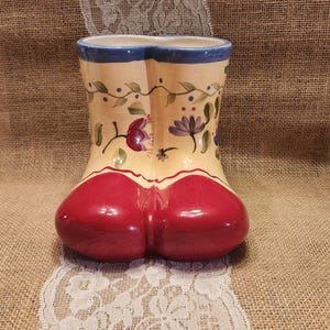May include: A ceramic boot-shaped vase featuring a glossy red base and a cream-colored upper section. The upper part is decorated with hand-painted floral motifs in shades of purple, red, and green, finished with a blue rim around the top opening.