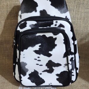 May include: A white and black cow print backpack with a front zippered pocket and black trim. The bag has a rounded shape and a single shoulder strap. The background is a neutral tan color.