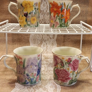 May include: Four ceramic mugs with gold-trimmed rims and handles, each featuring distinct floral illustrations. The top left mug displays yellow daffodils, the top right shows orange lilies, the bottom left features purple irises, and the bottom right depicts pink roses.
