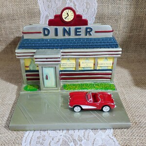 May include: A decorative diner figurine featuring a red convertible car parked in front. The building has a blue roof, a clock at the top, and signs reading DINER, TODAY'S SPECIAL, BURGERS & SODA, and LUNCH TO GO. The structure is painted in shades of gray, red, and blue.
