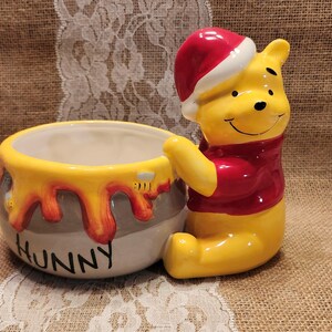 May include: A ceramic bowl shaped like a honey pot with the word HUNNY printed on the front in black letters. The pot features yellow honey dripping down the sides and a yellow bear figure wearing a red shirt and a red Santa hat attached.
