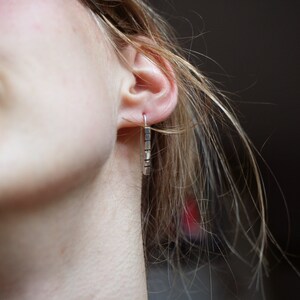 以下が含まれることがあります： 銀の棒状のイヤリングをした人の耳のクローズアップ。イヤリングは、複数の小さな長方形の棒でできています。