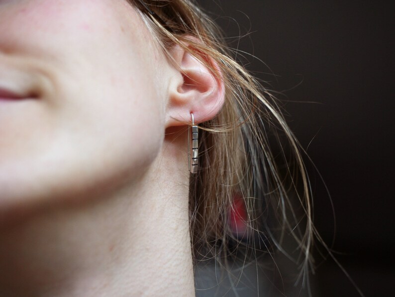 以下が含まれることがあります： 銀の棒状のイヤリングをした人の耳のクローズアップ。イヤリングは、複数の小さな長方形の棒でできています。