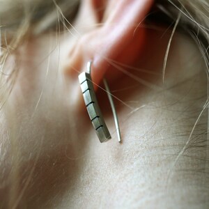 以下が含まれることがあります： 幾何学模様のシルバーバーイヤリング。イヤリングは人の耳にぶら下がっています。