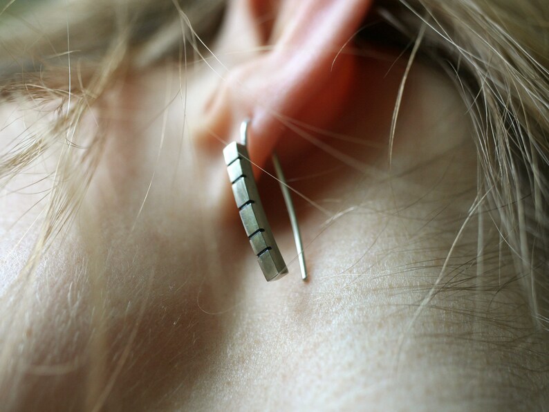 以下が含まれることがあります： 幾何学模様のシルバーバーイヤリング。イヤリングは人の耳にぶら下がっています。