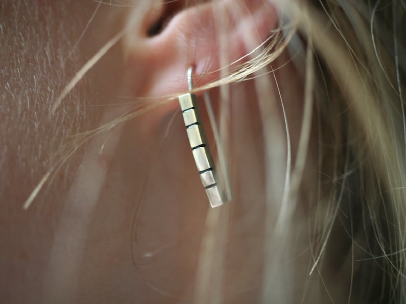 以下が含まれることがあります： テクスチャ加工された表面を持つシルバーバーのイヤリング。イヤリングは人の耳からぶら下がっています。