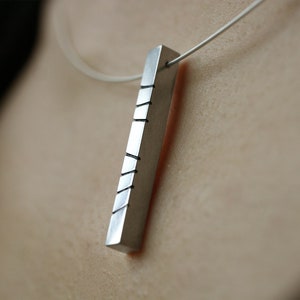 以下が含まれることがあります： 長い長方形のバーペンダントが付いたシルバーのペンダントネックレス。バーには、水平な線がいくつか刻まれています。