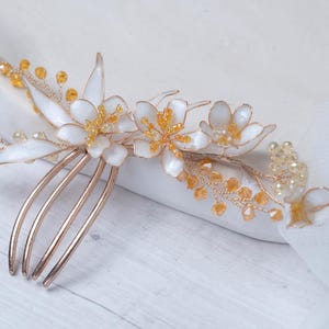Puede incluir: Una peineta con base de metal dorado adornada con pétalos de flores blancos y racimos de pequeñas cuentas de color ámbar translúcido. El diseño incluye delicadas ramas de alambre que se extienden desde las flores con diminutos cristales amarillos.