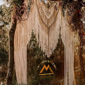 Könnte beinhalten: Ein Makramee-Hochzeitsbogen in Weiß und Beige. Der Bogen besteht aus dickem Seil und hat ein Fransen-Design. Der Bogen hängt an einem Baumzweig und ist von Grün umgeben. Der Text "MAYKNOTSSTUDIO Crafting with love" ist am unteren Rand des Bildes sichtbar.