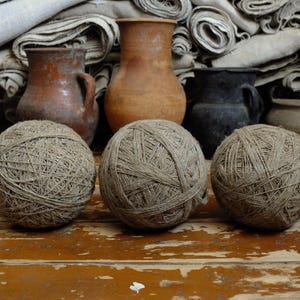 Op de afbeelding: Drie bollen touw liggen op een verweerd houten oppervlak. Achter hen staan ​​verschillende antieke keramische kruiken en een stapel gevouwen stof. De touwballen zijn lichtbruin en de kruiken in verschillende bruin- en zwarttinten.