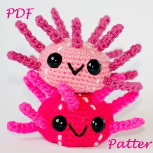 Puede incluir: Dos peluches de axolotl tejidos a crochet, uno rosa y otro rojo, apilados uno encima del otro. Los peluches tienen ojos negros, manchas blancas y una boca sonriente. El texto "PDF Pattern" es visible en la imagen.