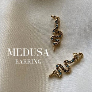 Könnte beinhalten: Ein Paar goldene Schlangenohrringe mit blauen Edelsteinen. Die Ohrringe liegen auf einem weißen Stoffhintergrund. Der Text "MEDUSA EARRING" ist unterhalb der Ohrringe sichtbar.