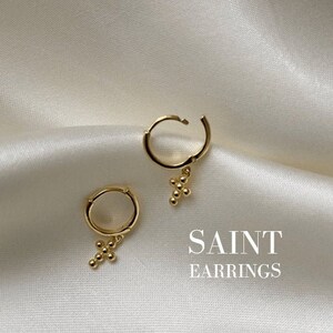 Könnte beinhalten: Ein Paar goldene Creolen mit einem kleinen Kreuzanhänger, der an jeder Creole hängt. Die Ohrringe liegen auf einem weißen Satinuntergrund. Der Text "SAINT EARRINGS" befindet sich unter den Ohrringen.