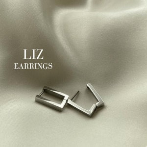 Könnte beinhalten: Silberfarbene quadratische Ohrringe auf cremefarbenem Satin-Hintergrund. Die Ohrringe haben eine geometrische Form, mit einem rechteckigen Bügel und einem Stift. Der Text "LIZ EARRINGS" ist weiß gedruckt.