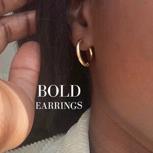 Könnte beinhalten: Nahaufnahme eines Ohrs mit einem Paar goldenen Creolen. Der Text "BOLD EARRINGS" steht unter den Ohrringen.