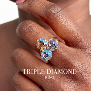 Könnte beinhalten: Ein goldener Ring mit drei schillernden, mehrfarbigen Edelsteinen. Der Ring heißt "Triple Diamond Ring".