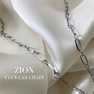 May include: A silver chain necklace with a simple, linked design. The chain is on a white fabric background. The text "ZION EYEWEAR CHAIN" is printed below the chain.