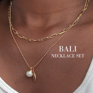 Könnte beinhalten: Ein goldener Halsketten-Set mit einer zarten Kette und einer größeren Kette mit einem Perlenanhänger. Das Set heißt "BALI NECKLACE SET".