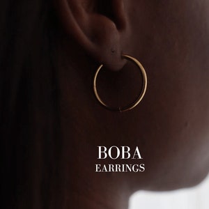 Könnte beinhalten: Ein Paar goldene Creolen. Die Ohrringe sind schlicht und elegant und eignen sich perfekt als Geschenk für jeden Anlass. Der Text "BOBA EARRINGS" ist unter den Ohrringen zu sehen.