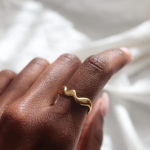 Könnte beinhalten: Ein goldfarbener Ring mit einem wellenförmigen Design. Der Ring befindet sich an einem Finger, vor einem weichen, weißen Hintergrund. Das Design des Rings ist fließend und organisch, mit poliertem Finish. Der Ring ist ein Schmuckstück.