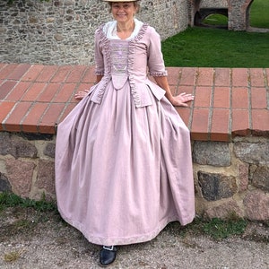 May include: A woman wearing a pink, long-sleeved dress with a white lace trim and a straw hat with a feather. She is standing in front of a stone wall with a brick walkway.