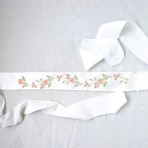 May include: A long, white fabric sash featuring a central embroidered floral vine pattern. The design includes small pink and orange flowers with green stems and leaves. The sash has long, unhemmed, flowing fabric ties extending from both ends.