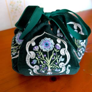 May include: A dark green velvet drawstring bag with embroidered floral designs in shades of blue, purple, and silver. The bag has a green velvet ribbon closure.