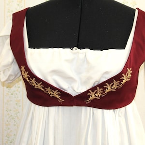 May include: A white dress featuring a deep square neckline and a dark red bodice overlay. The bodice is decorated with gold-coloured floral embroidery along the lower edge. The dress has short, puffed sleeves and a gathered skirt with a high waistline.