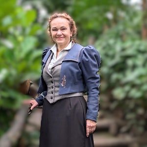 May include: A woman wearing a blue jacket with a brown and white plaid vest, a black skirt, and a white blouse. She is standing in front of a green background.