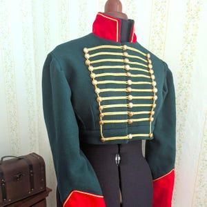 May include: A dark green cropped jacket with gold trim and buttons, red collar and cuffs. The jacket is displayed on a mannequin. The jacket has a military style with multiple rows of gold braid and decorative buttons.