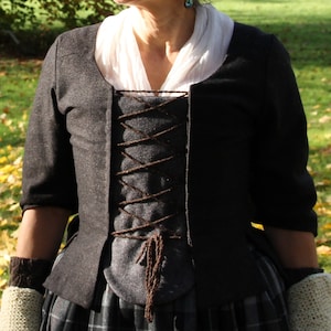 以下が含まれることがあります： 濃いグレーのジャケットで、フロントは茶色のレースアップになっている。ジャケットはフィットしたボディと少し広がったスカートになっている。ジャケットは厚くて質感のある生地で作られている。