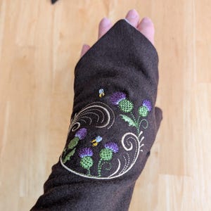 Op de afbeelding: Een donkerbruine vingerloze handschoen met een geborduurd ontwerp van paarse en groene distelbloemen, krullende crèmekleurige ranken en twee kleine gele en blauwe bijen. Het borduurwerk is gecentreerd op de rug van de hand.